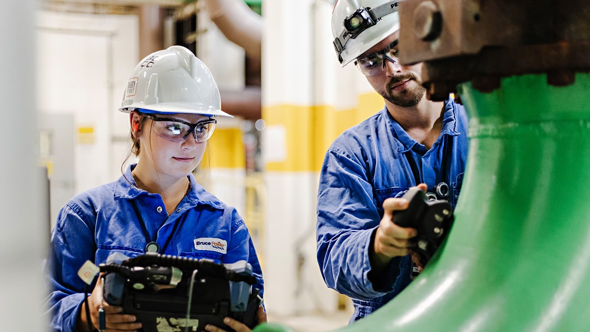 Woman and man, wearing a hardhat and safety goggles, inspecting equipment.