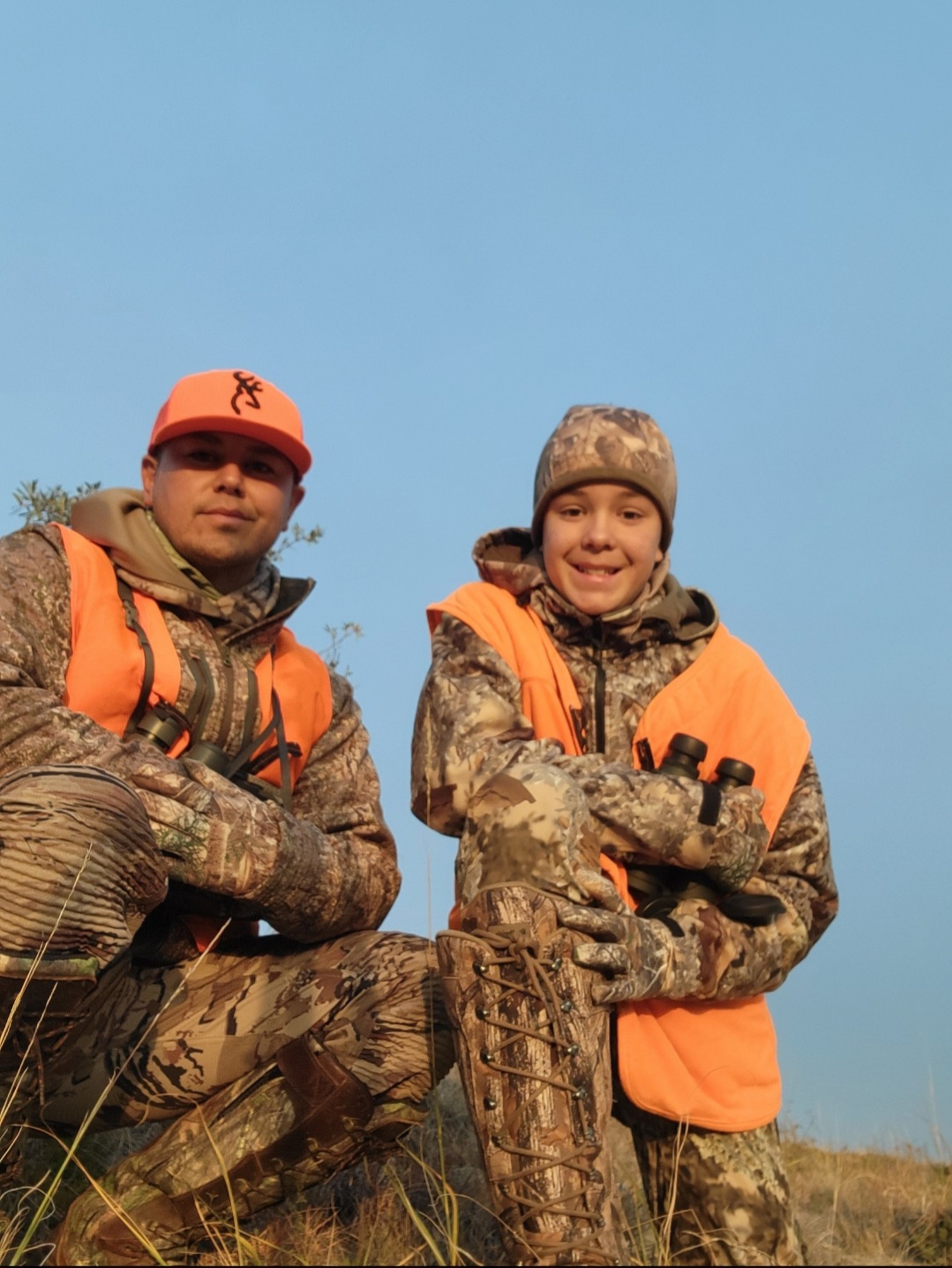 A man and a child, both wearing orange hunting vests and camouflage gear, are kneeling on grassy terrain.