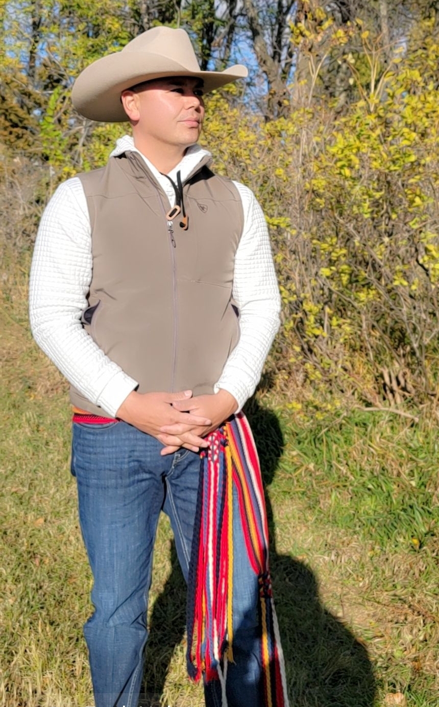 A man stands outdoors, wearing a hat and vest, holding a colorful woven sash.